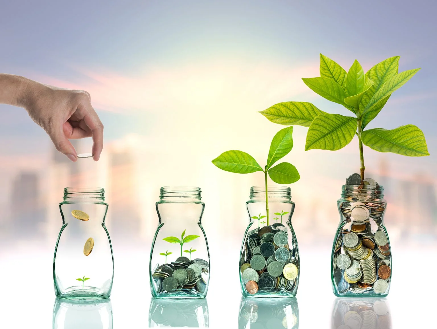 Hand putting mix coins and seed into 4 clear bottles that are increasing in number of coins and size of plants