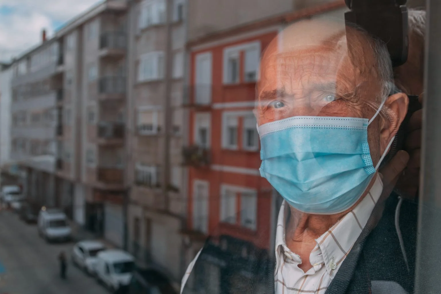 senior with face mask confined at home looking out the window