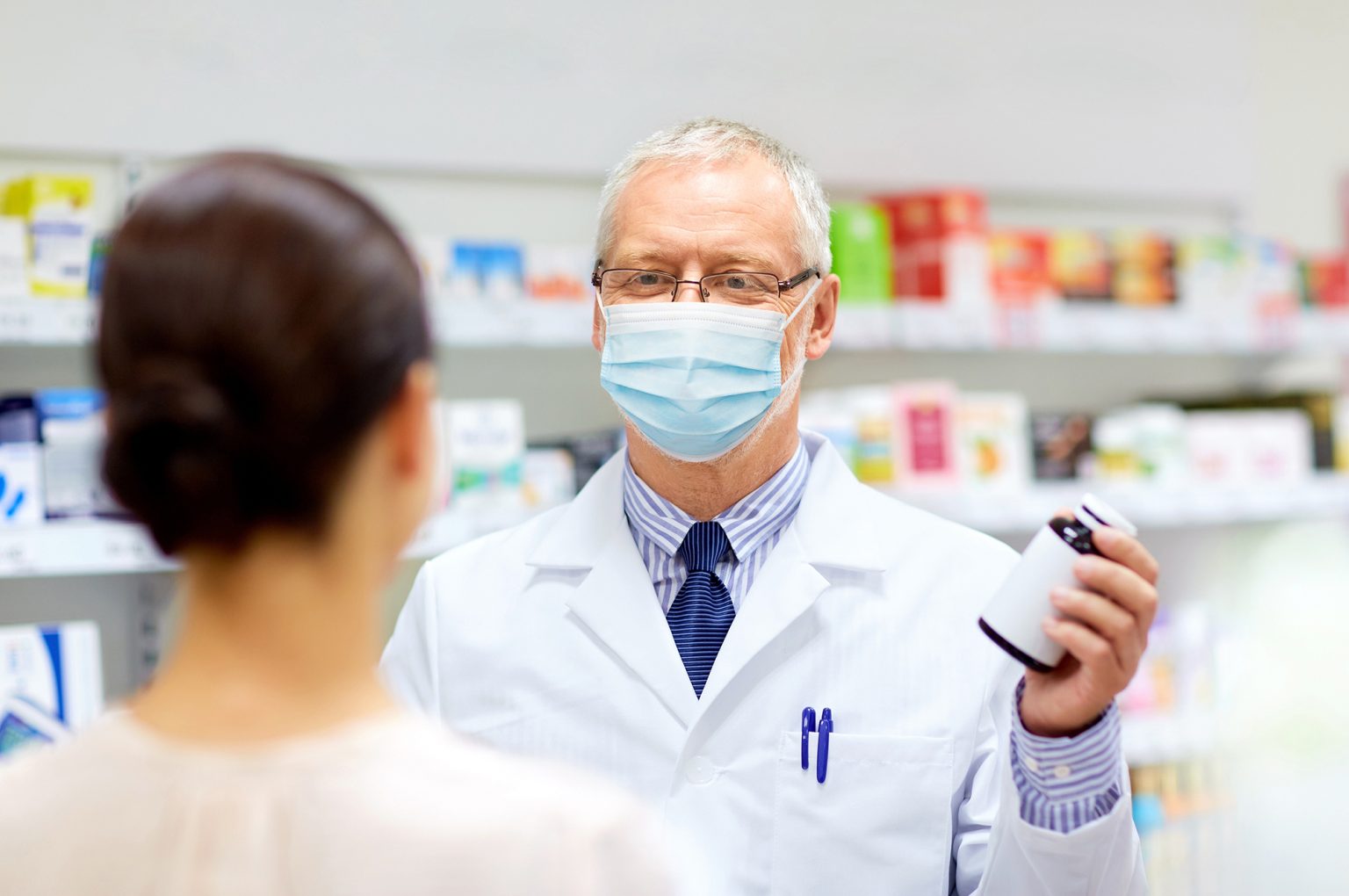 Older pharmacist wearing a mask giving prescription to female customer