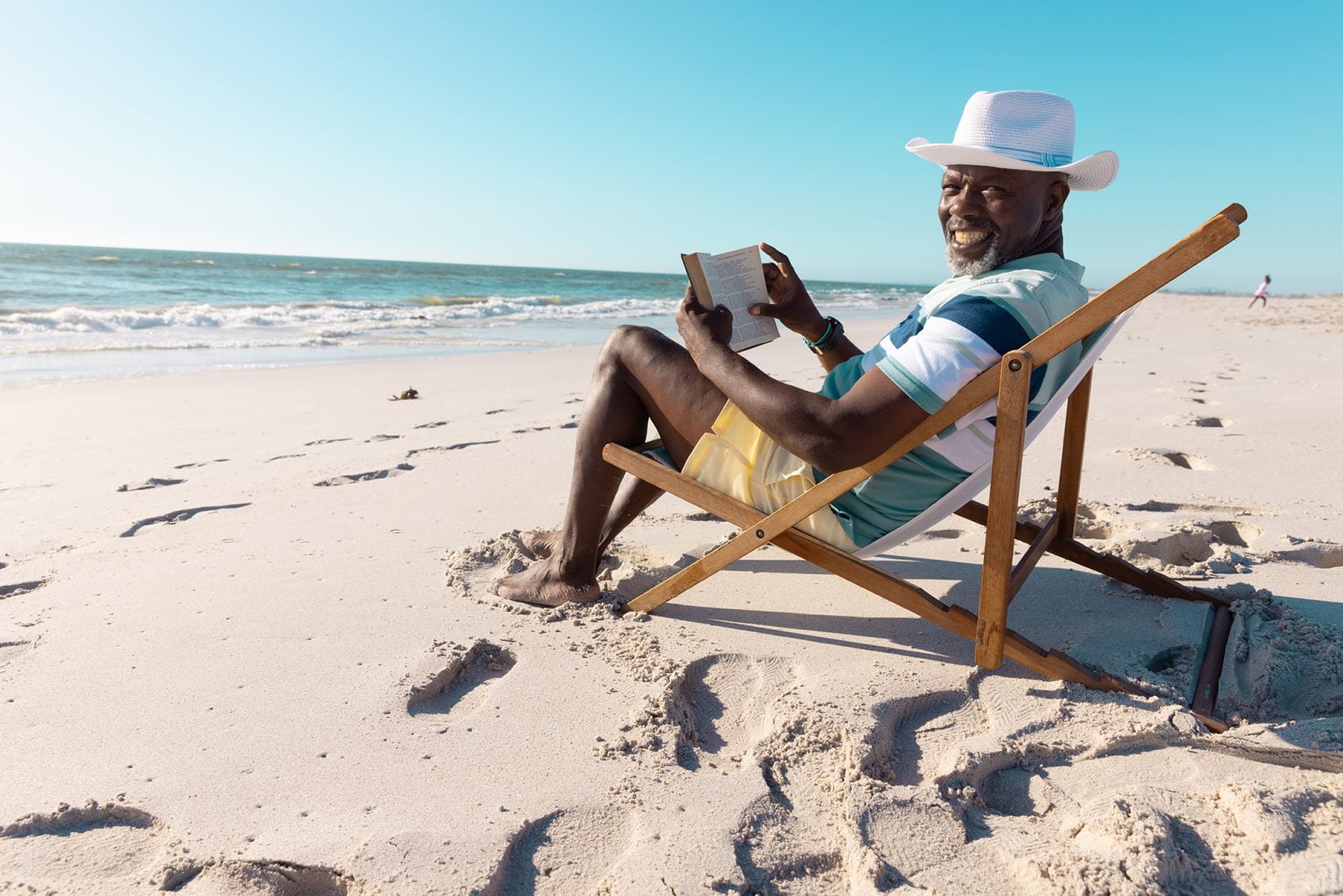 senior man wearing hat reading book on deckchair at beach under clear sky