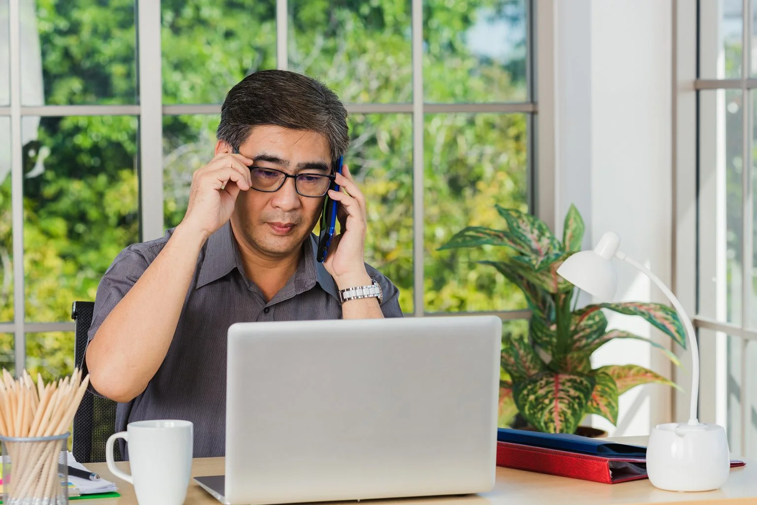 Senior man sitting at a table using a laptop and talking on a cellphone