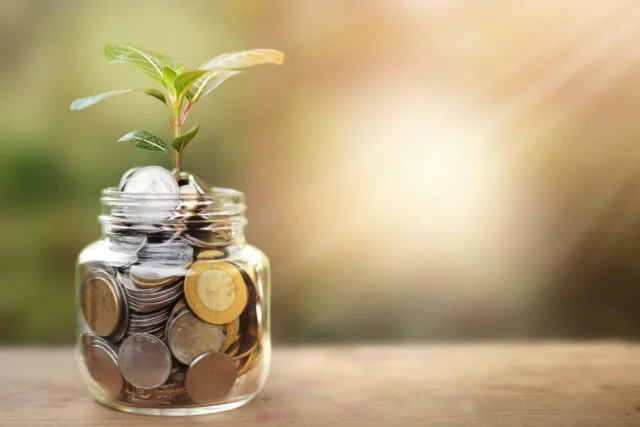 A glass jar full of coins and plant growing through it with some coins and plant leaves.