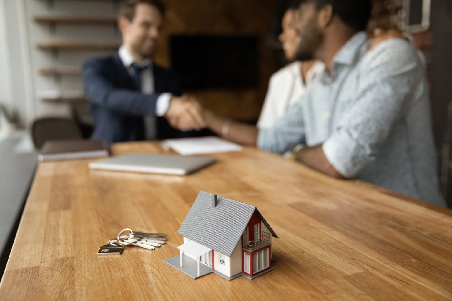 Couple shaking hands with realtor with a model house and keys in foreground