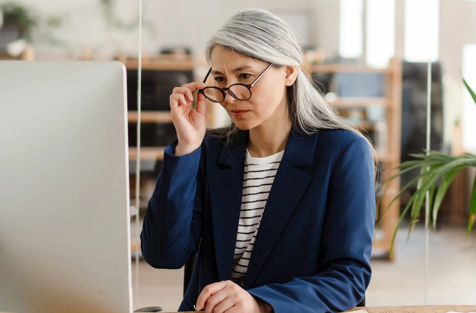 Older woman sitting at a table in front of the monitor adjusting glasses with her hand in the office