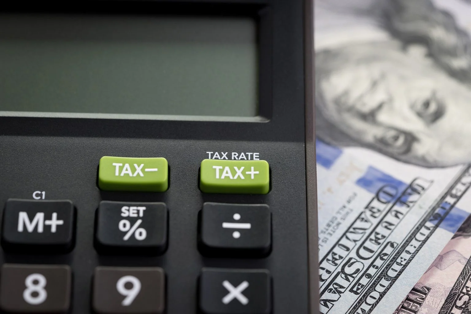 Closed up shot of TAX minus / plus buttons with text TAX RATE on calculator with background of blurred US Dollar banknotes