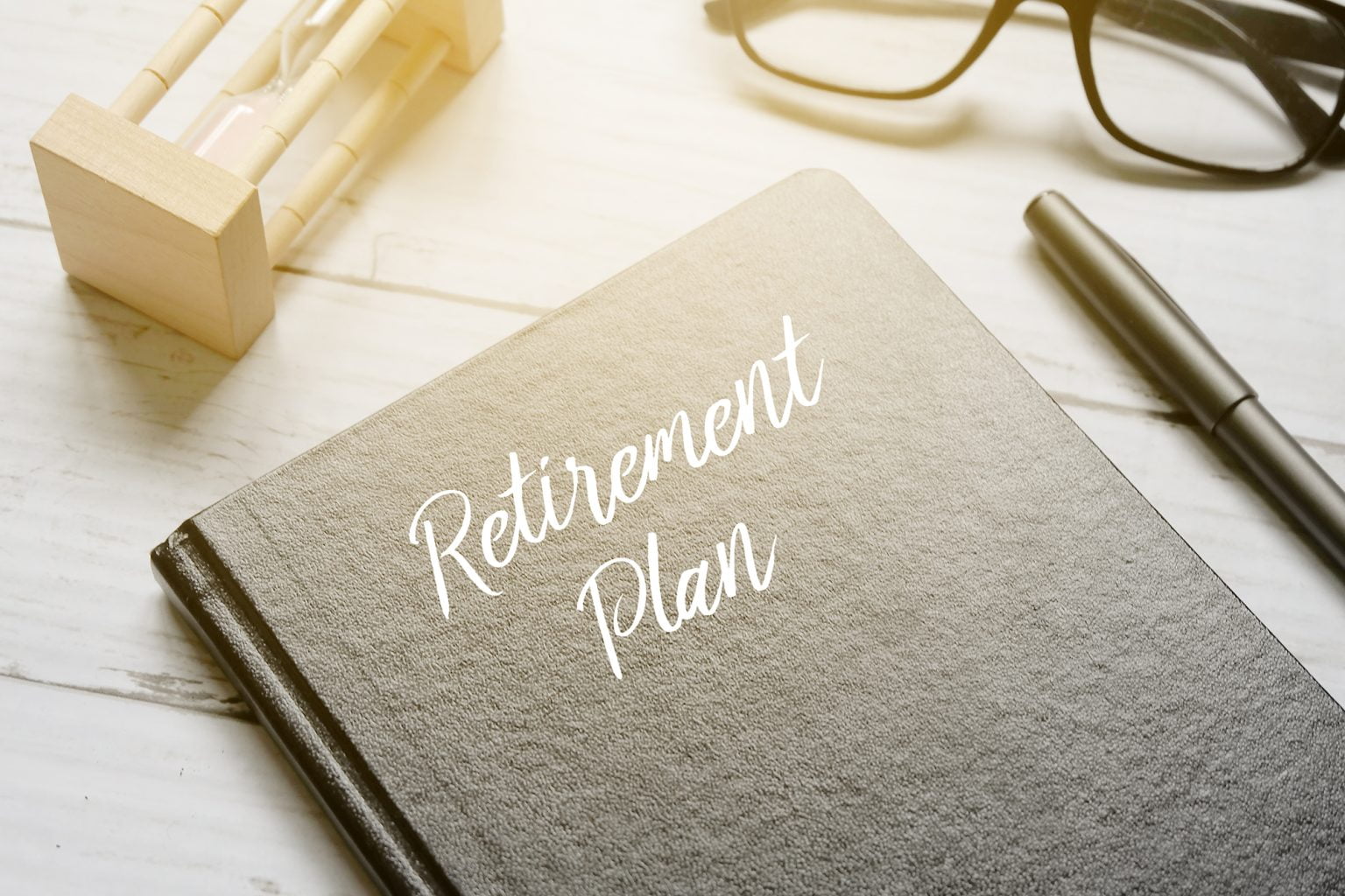 Hourglass, eyeglass, pen and notebook written with RETIREMENT PLAN on white wooden background with sun flare
