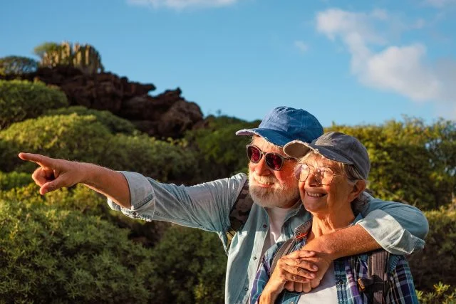 Smiling old retirees in hat and casual clothes among green bushes and blue sky
