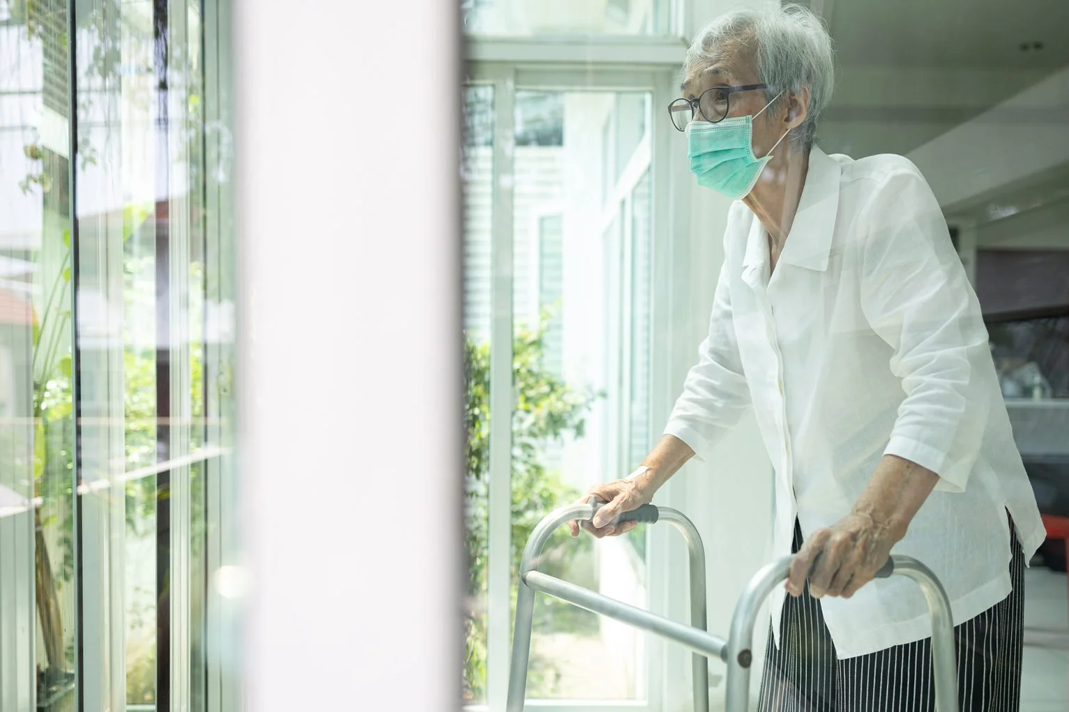 senior woman is standing alone wiuth walker in front of a window