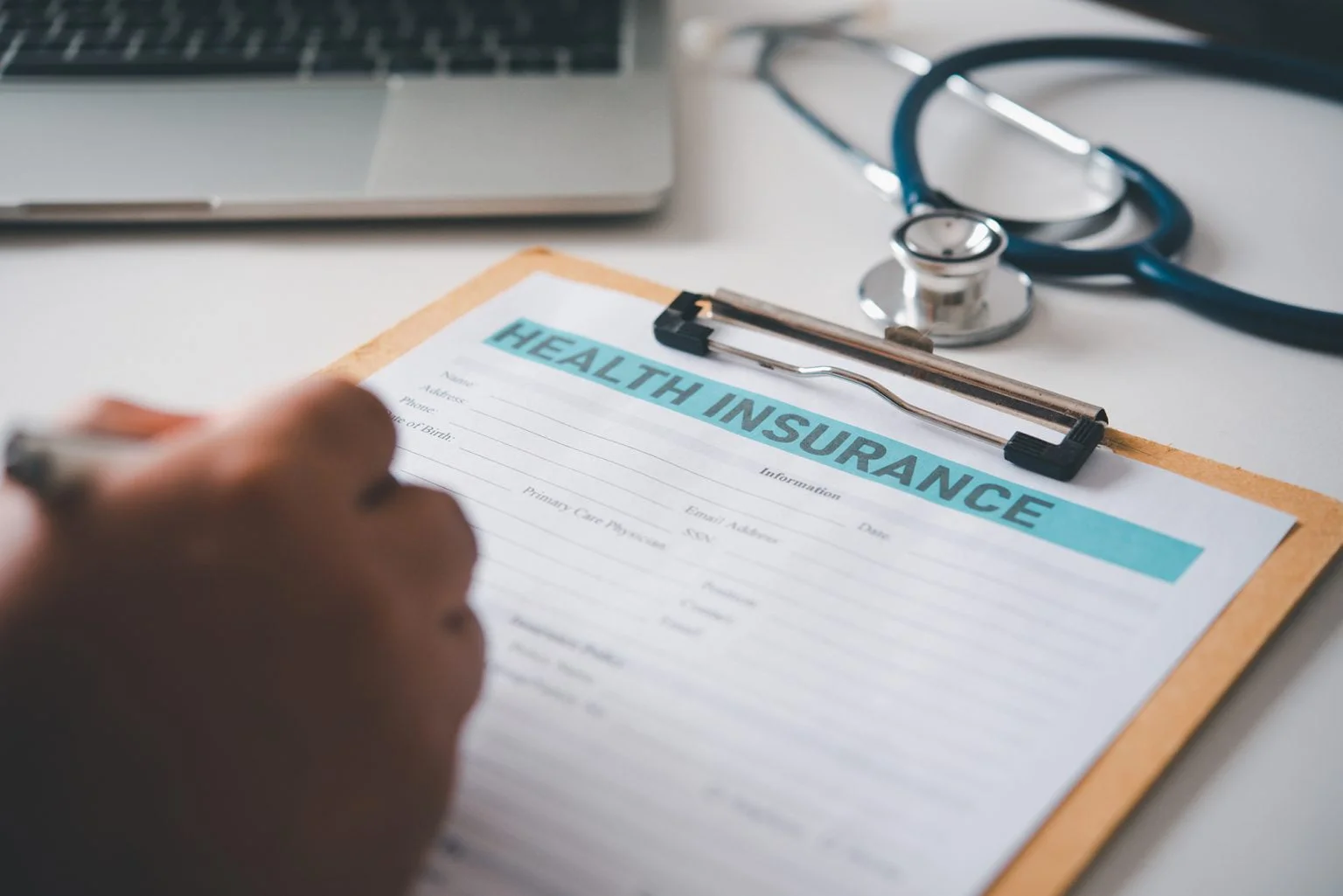 Hand fillin gout a health inaurance form on a clipboard with. a stethoscope and laptop in the background