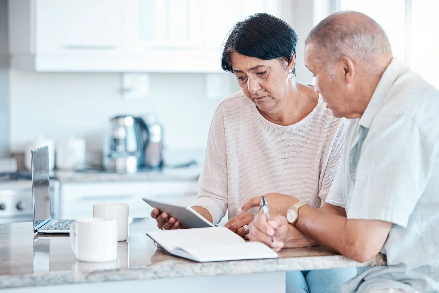Senior couple looking at a tablet, laptop and open notebook at a kitchen counter