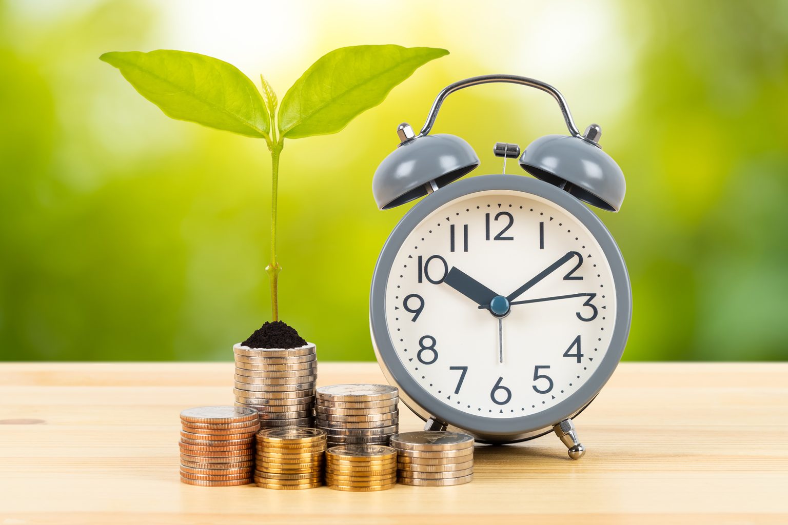 Coin stack with growing leaves and alarm clock on wooden desk on green tree background, time for saving concept