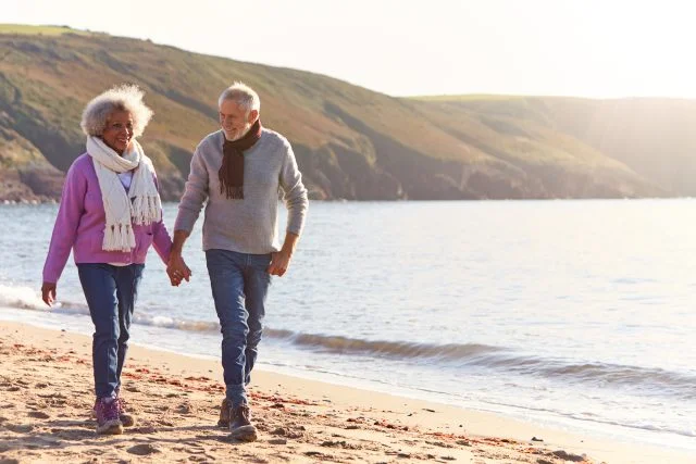 Loving Retired Couple Holding Hands As They Walk Along Shoreline On Winter Beach Vacation