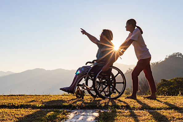 Disabled handicapped young man in wheelchair walking with his care helper in sunset.