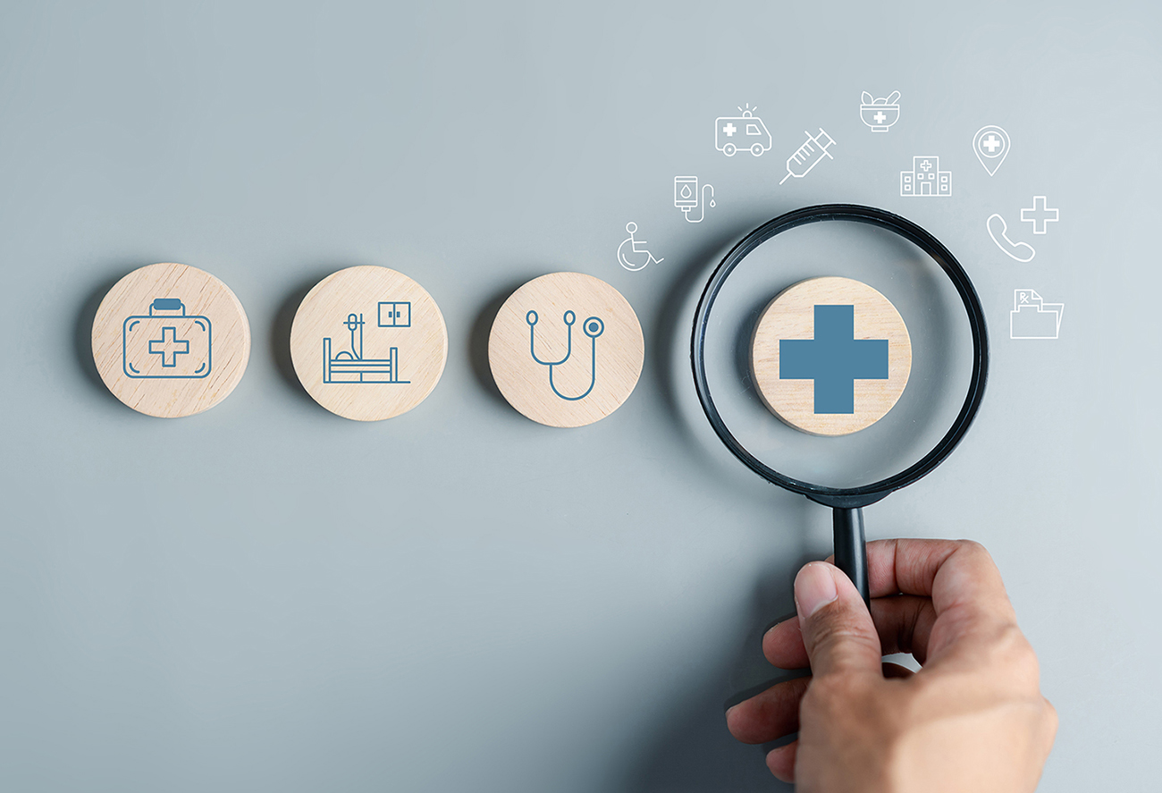 Close up of a hand holding a magnifying glass over wooden circles with health insurance icons