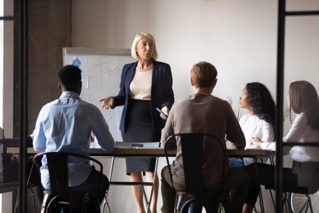 Older woman presenting to colleagues in front of a whiteboard