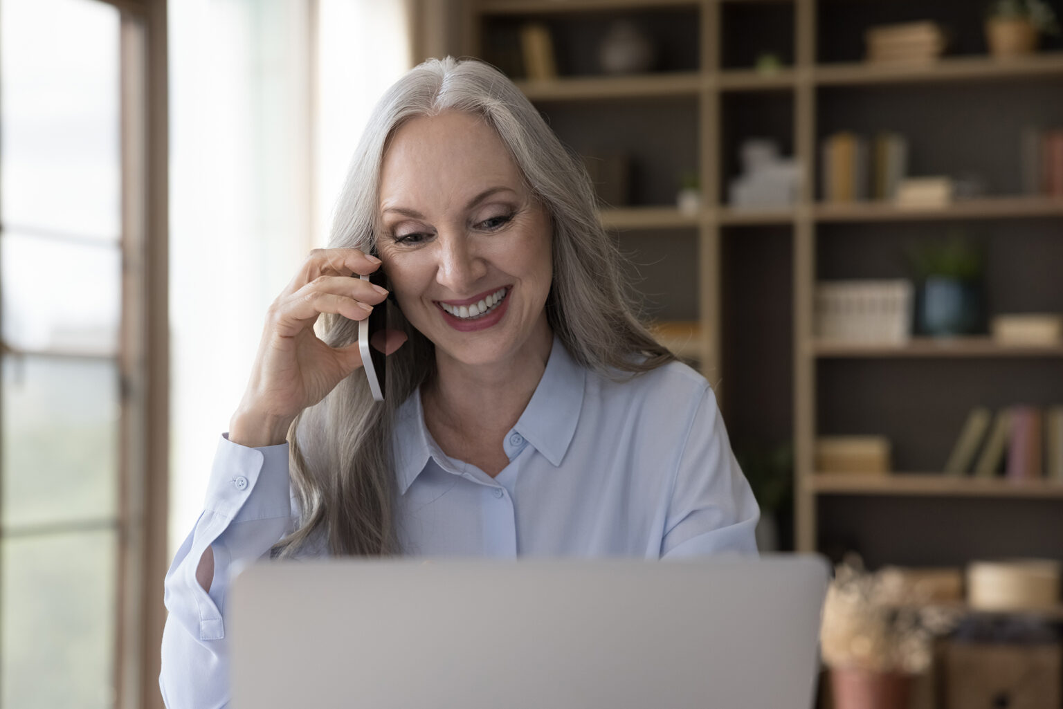 Older woman smiling talking on a cellphone in front of a computer at home