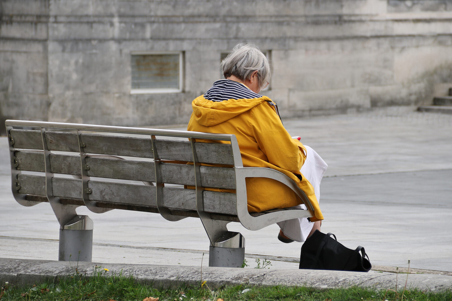 View of an older woman's back sitting on a city bench wearing a yellow coat