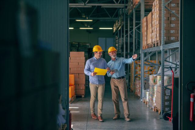 CEO going around warehouse with supervisor and talking analyzing sale statistics. Younger man holding folder with data while older one holding tablet and pointing at boxes. Both having yellow helmets.