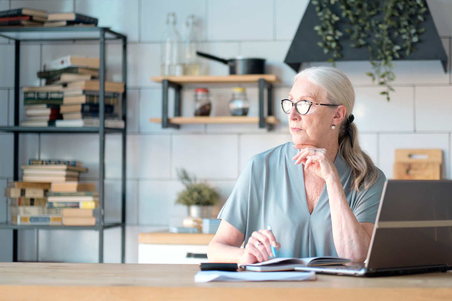 Senior woman using laptop for websurfing in her kitchen. The concept of senior employment, social security. Mature lady sitting at work typing a notebook computer in an home office