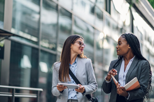 Two women walking outside one holding a tablet one holding a book looking at each other