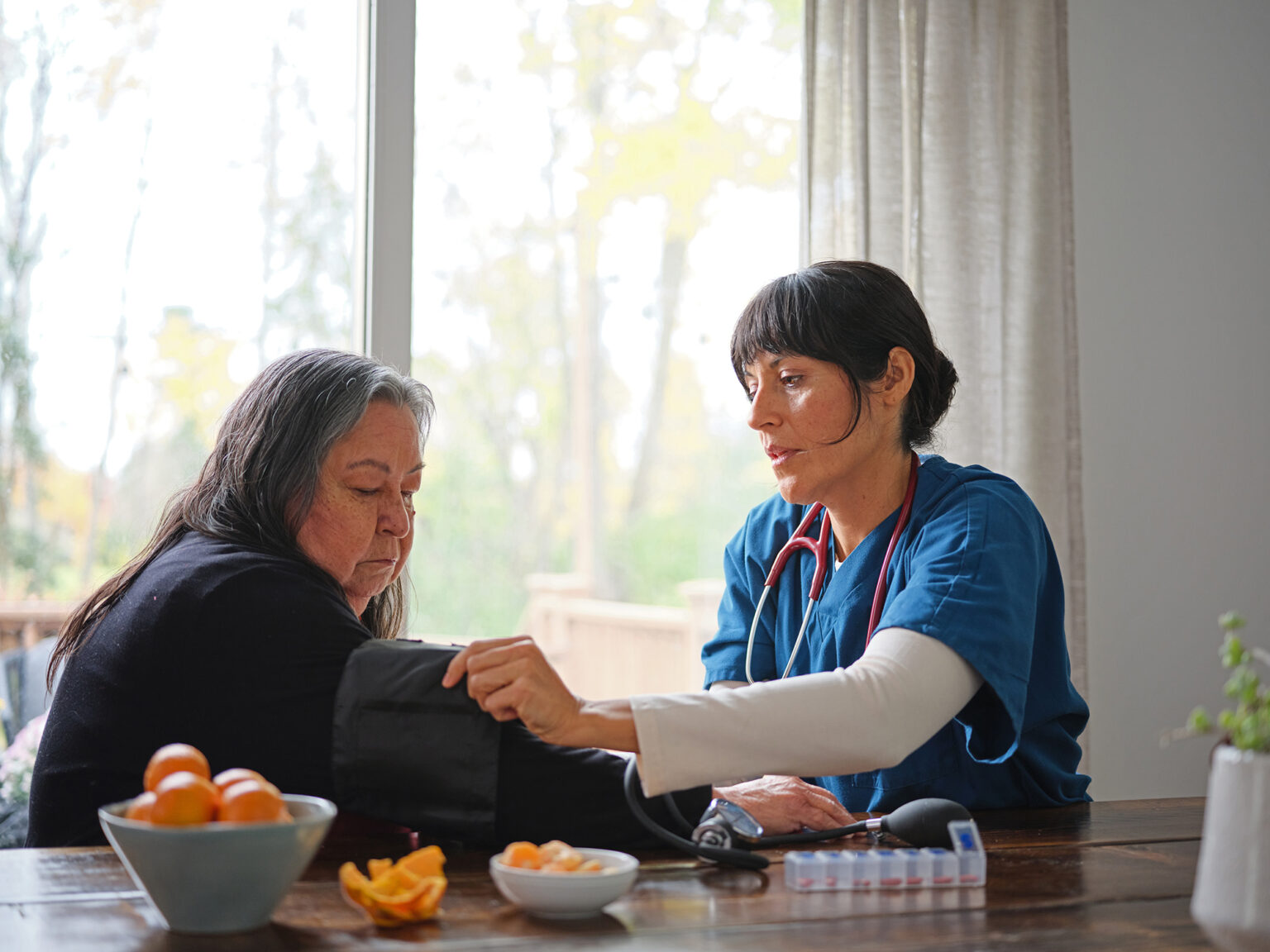 A nurse fits a blood pressure monitor on a senior woman at a kitchen table