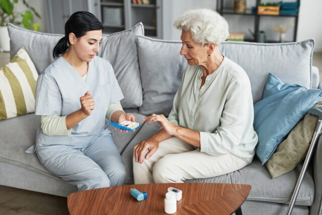 Portrait of a young nurse showing pill box to a senior woman sitting on a couch at home